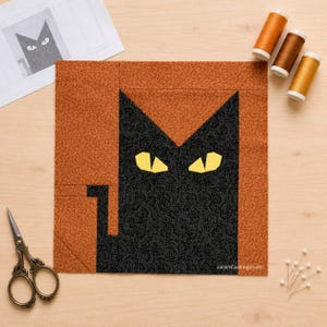 Puede incluir: Un bloque de edredón cuadrado con la silueta de un gato negro con ojos amarillos sobre un fondo naranja texturizado. El gato tiene orejas puntiagudas y una cola pequeña. Se ven tres carretes de hilo, tijeras de costura y un patrón de papel.