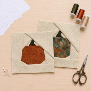 May include: Two square fabric quilt blocks featuring a central pumpkin shape pieced with orange and teal patterned fabric, surrounded by cream-colored paisley fabric. Includes three spools of thread, sewing pins, and ornate metal scissors.