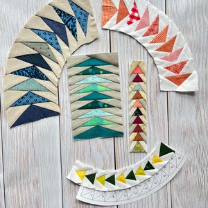 Pode incluir: Sortido de peças de tecido em várias formas e cores, dispostas sobre uma superfície de madeira clara. As peças incluem segmentos curvos com triângulos azuis, laranja e rosa, juntamente com tiras retangulares e estreitas. Também está presente um modelo de papel com números.