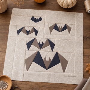 Könnte beinhalten: Ein quadratischer Stoff-Quiltblock mit beigem Hintergrund und kleinem weißen Blumenmuster. Der Block zeigt fünf zusammengesetzte Fledermausformen in verschiedenen Größen, gefertigt aus dunkelblauen und hellbraunen Stoffstücken. Der Text Jane's Quilting Room ist in der unteren rechten Ecke aufgedruckt.