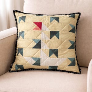 May include: Square cushion with a geometric pattern. The cushion has a pale yellow background with a quilted design of white, teal, and red shapes. It is finished with a black border.