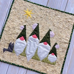 Puede incluir: Un camino de mesa navideño acolchado con cuatro figuras de gnomos con barbas blancas, narices rosas y sombreros verdes. Una estrella amarilla está en la esquina superior izquierda. El camino de mesa tiene un fondo beige con un borde verde.