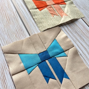 以下が含まれることがあります： 手作りのリボンデザインの布ブロックが2つ。1つのブロックは、ベージュの背景にオレンジ色のリボンと繊細な模様が特徴です。もう1つのブロックは、ベージュの背景に青いリボンがあります。ブロックは明るい木製の表面に配置されています。