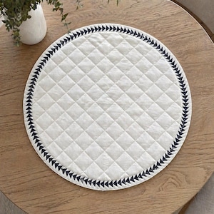 May include: A round, white quilted placemat with a navy blue triangle border. The placemat is on a light brown wooden table. A small white vase with green foliage is in the background.