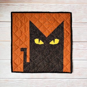 May include: A square quilted wall hanging featuring a black cat face with yellow eyes against an orange background. The cat's face is centered, with a black border and a quilted diamond pattern.