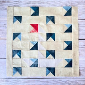 Peut inclure: Une courtepointe faite à la main avec un motif répétitif de triangles bleu foncé, bleu clair, blancs et rouges sur un fond crème. La courtepointe a une bordure crème et est exposée sur une surface en bois clair.