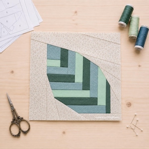May include: A square quilt block featuring a central leaf-shaped motif constructed from horizontal fabric strips in shades of dark green, sage, and light mint, surrounded by a speckled beige border, with sewing scissors and thread spools.