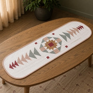 May include: An oval-shaped quilted table runner featuring a white fabric base with a grid pattern. The design includes a central star motif in shades of brown, red, and sage green, flanked by rows of triangular shapes in red, pink, and sage green.