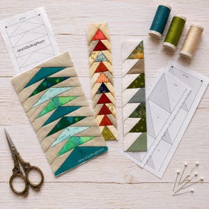 May include: A quilting project featuring three fabric strips with triangular patterns in shades of green, red, and earth tones. Includes paper templates labeled Jane'sQuiltingRoom with 2.5 inch markings, three spools of thread, and sewing pins.