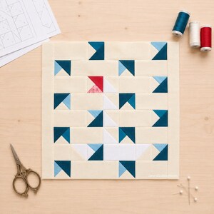 May include: A square quilt block featuring a central white swan motif with a red beak, constructed from cream, white, dark teal, and light blue fabric pieces. The design is surrounded by a cream border, with sewing supplies visible nearby.