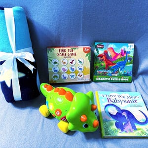May include: A gift set featuring a rolled-up blue blanket tied with a white ribbon, a green dinosaur toy, a "Find the Same Game" box, a "Magnetic Puzzle Book", and a book titled "I Love You More, Babysaur". The items are arranged on a blue surface.