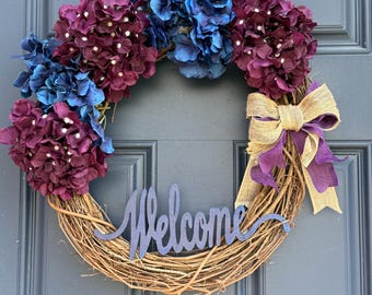 May include: A grapevine wreath decorated with purple and blue hydrangea flowers and a burlap bow. The wreath has a wooden sign that says "Welcome".