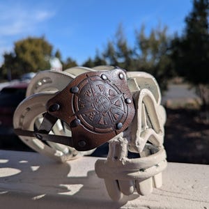 Könnte beinhalten: Eine weiße Metallskulptur eines Schädels mit einem braunen Lederaufnäher auf der Stirn. Der Lederaufnäher hat ein Kompassdesign. Der Schädel besteht aus verbundenen Hufeisen.