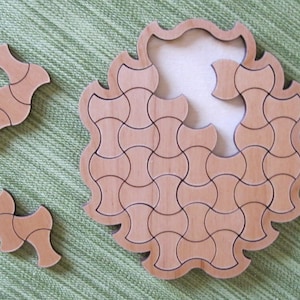 Puede incluir: Un rompecabezas de madera circular con una pieza que falta. El rompecabezas está hecho de pequeñas piezas curvas que encajan para crear un diseño más grande. La pieza que falta se muestra por separado.