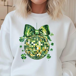 Op de afbeelding: Wit sweatshirt met een groene en gouden discobal met een groene strik erop. De discobal is omringd door groene klavertjes.