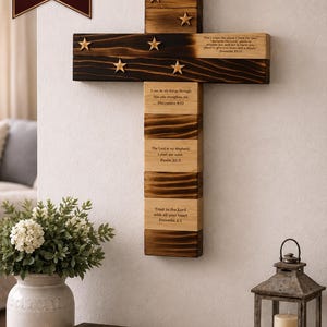 May include: A wall-mounted wooden cross featuring a dark charred finish on the upper sections and natural wood on the lower parts. It is decorated with small wooden stars and engraved with Bible verses including Jeremiah 29:11 and Psalm 23:1.