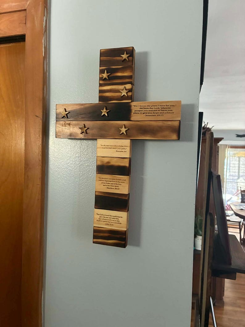 May include: A wall-mounted wooden cross with a scorched finish, featuring seven attached wooden stars. The cross displays printed Bible verses including Jeremiah 29:11, Proverbs 3:6, Matthew 28:19, and John 3:16 on the horizontal and vertical segments.