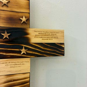 May include: A wooden cross wall hanging featuring burnt wood edges and attached wooden stars. The surface displays printed Bible verses including Jeremiah 29:11, Philippians 4:13, and Matthew 28:19, with visible wood grain and charred dark brown accents.