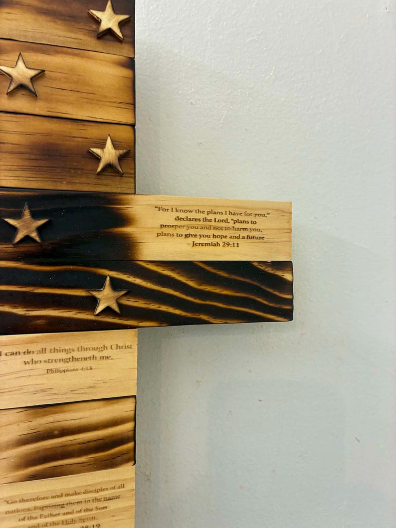 May include: A wooden cross wall hanging featuring burnt wood edges and attached wooden stars. The surface displays printed Bible verses including Jeremiah 29:11, Philippians 4:13, and Matthew 28:19, with visible wood grain and charred dark brown accents.