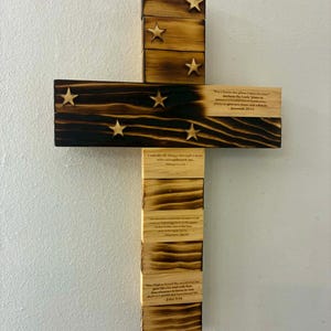 May include: A wooden cross wall decoration featuring a burnt wood finish on the upper section with six attached wooden stars. The lower section is light-colored wood with printed Bible verses including Jeremiah 29:11, Philippians 4:13, Matthew 28:19, and John 3:16.