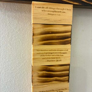 May include: A vertical wooden wall hanging featuring alternating blocks of light natural wood and scorched dark wood. The light blocks are printed with Bible verses: Philippians 4:13, Matthew 28:19, and John 3:16. A dark wooden crossbar with stars sits above.
