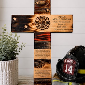 May include: A scorched wooden cross featuring a firefighter emblem, the text "CAPTAIN MICHAEL THOMPSON Dayton Fire Department Engine 14 1998-2026 28 Years of Service," and two Bible verses from Isaiah 43:2 and Joshua 1:9 engraved on the surface.