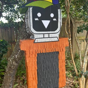 May include: A piñata in the shape of a computer monitor, complete with a colourful baseball cap. The monitor features a black screen with white details and a silver border. The base is orange and black, made of fringed paper.