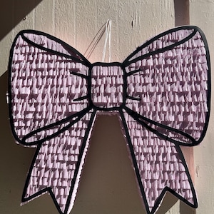 Op de afbeelding: Een grote, roze strikvormige piñata met een zwarte omtrek. De strik is gemaakt van getextureerd, gefranjerd papier. Er is een wit touwtje bevestigd om op te hangen. De piñata staat tegen een lichtroze muur.