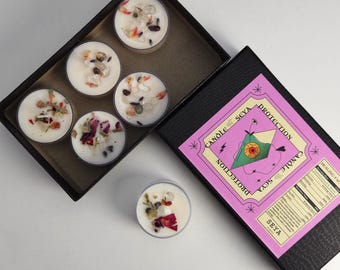 May include: A set of six white tea light candles in a black box. Each candle is decorated with dried flowers and crystals. The box has a pink label with the words "Protection Candle" and "Seya".