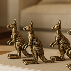 May include: Four bronze-colored kangaroo figurines are arranged on a stack of books. The kangaroos are in a standing pose, with their tails curled. The figurines have a detailed, textured surface. The books are light beige with a neutral background.