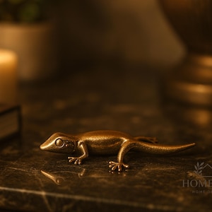May include: A bronze-colored metal lizard figurine. The lizard is detailed with visible legs, a tail, and a head with eyes. The figurine is placed on a dark, textured surface. The background includes a lit candle, a book, and a decorative lamp.
