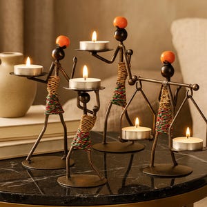 May include: Decorative metal candle holders in the shape of stylized figures. Each figure holds a lit tea light candle. The figures have orange spheres for heads and are adorned with wrapped, colorful accents. The candle holders are on a dark, patterned surface.