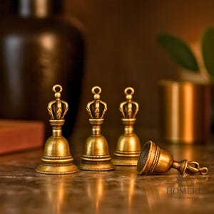 May include: Four antique brass bells with crown-shaped handles. Three bells stand upright, while one rests on its side. The bells have a detailed, textured surface and a warm, golden hue, suggesting a vintage or decorative item.