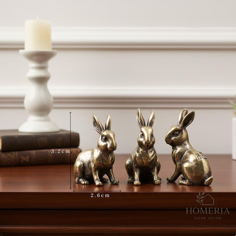 May include: Three bronze-colored rabbit figurines, each approximately 2.6 cm long and 3.2 cm tall, are displayed on a dark wooden surface. The rabbits are in different poses. A white candle holder and books are in the background.