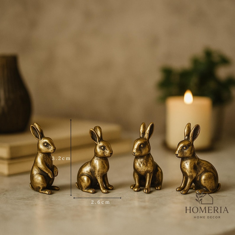 May include: Four small, bronze-colored rabbit figurines. One rabbit is standing, while the others are seated. The figurines are approximately 3.2 cm tall and 2.6 cm wide. The background includes a candle and the text "HOMERIA HOME DECOR."
