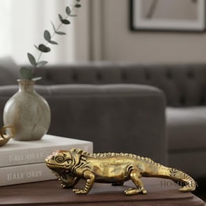 May include: A detailed, golden-colored iguana sculpture is displayed on a dark wooden surface. The sculpture is positioned in front of a small, light-colored vase with a sprig of greenery. The word "HOMERIA" is visible on the sculpture.