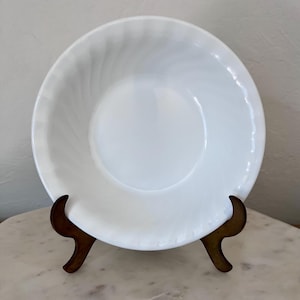 May include: A round, white milk glass bowl featuring a subtle, swirling ribbed pattern along the wide rim. The bowl has a smooth, glossy finish and a deep, circular center, presented on a dark brown, curved metal display stand for viewing.