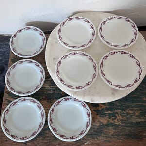 May include: A set of seven white milk glass plates featuring a repeating maroon wave pattern along the rim. The plates are circular with a smooth finish and a consistent design, arranged in a group on a circular white marble surface.