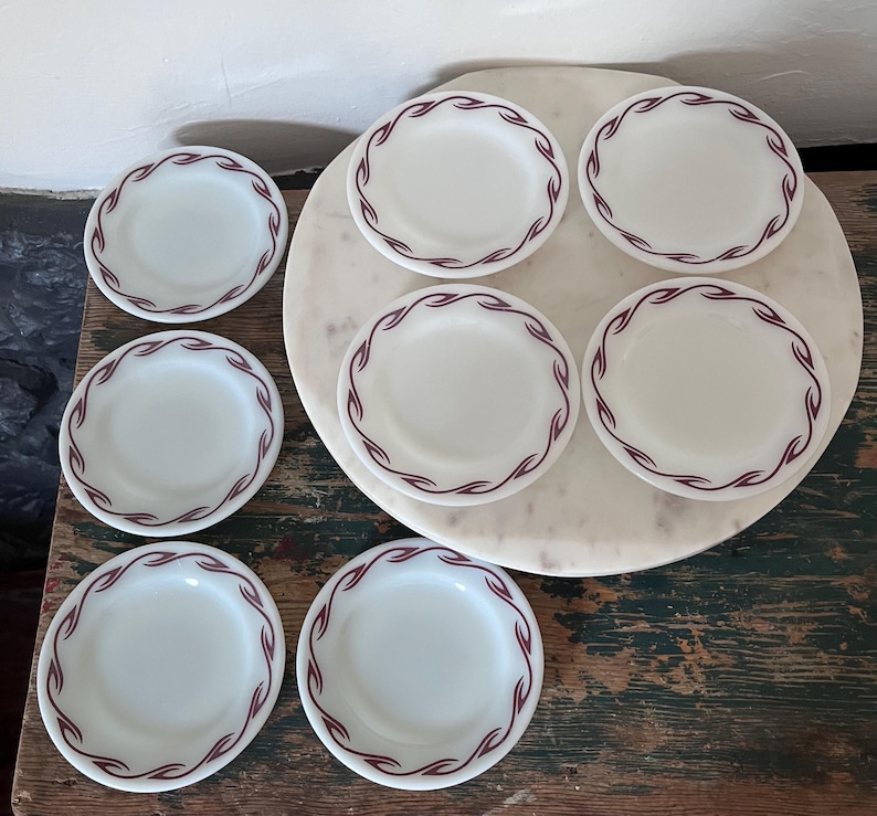 Puede incluir: Un juego de siete platos de vidrio lechoso blanco con un patr&oacute;n repetitivo de ondas color granate a lo largo del borde. Los platos son circulares, tienen un acabado liso y un dise&ntilde;o consistente, dispuestos sobre una superficie de m&aacute;rmol blanco.