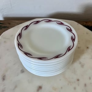 Puede incluir: Una pila de platos peque&ntilde;os y redondos de vidrio lechoso blanco con un patr&oacute;n repetitivo de color granate en forma de llama a lo largo del borde. Los platos son opacos con un acabado liso y brillante, y el dise&ntilde;o consiste en formas curvas estilizadas.