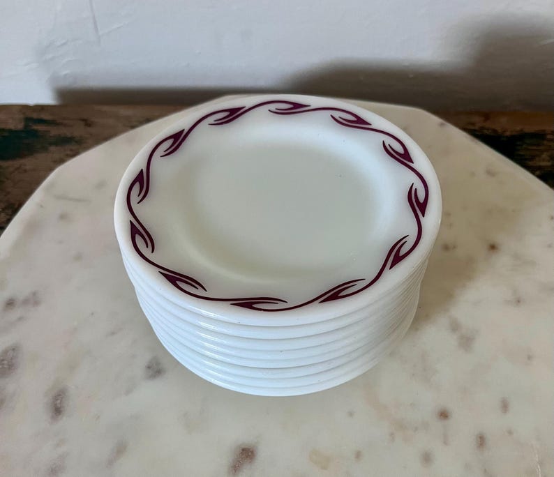 Puede incluir: Una pila de platos peque&ntilde;os y redondos de vidrio lechoso blanco con un patr&oacute;n repetitivo de color granate en forma de llama a lo largo del borde. Los platos son opacos con un acabado liso y brillante, y el dise&ntilde;o consiste en formas curvas estilizadas.