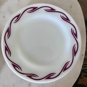 Puede incluir: Un plato redondo de vidrio lechoso blanco que presenta un patr&oacute;n de borde continuo de formas estilizadas de hojas o llamas de color p&uacute;rpura oscuro. El plato tiene un acabado liso y brillante y un dise&ntilde;o circular simple.
