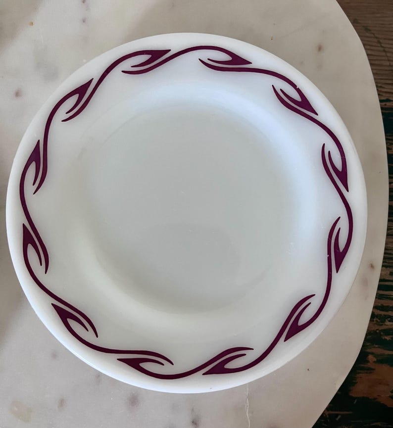 Puede incluir: Un plato redondo de vidrio lechoso blanco que presenta un patr&oacute;n de borde continuo de formas estilizadas de hojas o llamas de color p&uacute;rpura oscuro. El plato tiene un acabado liso y brillante y un dise&ntilde;o circular simple.