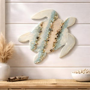 May include: A decorative turtle wall art piece. The turtle is made of a light cream-colored material with a blue and beige center, adorned with small seashells. The art piece is mounted on a white, horizontal-slat wall.