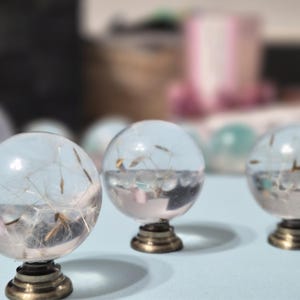 May include: Three clear glass orbs with dandelion seeds encased inside, resting on ornate brass bases. The orbs are arranged on a light blue surface, with a blurred background. The orbs are decorative.