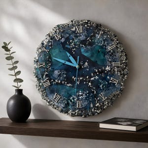May include: A round wall clock with a blue and teal resin face, accented with silver-colored embellishments. The clock features silver Roman numerals and light blue hands. The clock is displayed on a wall above a dark wooden shelf.