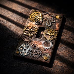 May include: A rectangular, steampunk-style light switch cover. The cover is a rusty brown with various sizes of gold and silver gears and cogs affixed to the surface. The cover has a keyhole and is set against a textured background.