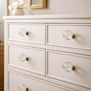 May include: A white dresser with four drawers, each adorned with a clear, spherical glass knob and a brass base. The knobs add a touch of elegance to the otherwise simple design. The dresser is set against a neutral background.