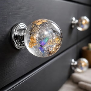 Op de afbeelding: Transparante glazen kastknoppen met een zilveren basis en een decoratief interieur. De binnenkant van de knoppen heeft gouden vlokken, zilveren glitter en iriserende accenten. De knoppen zijn gemonteerd op een zwarte kast.