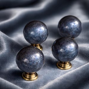 May include: Four spherical cabinet knobs with a dark blue, speckled finish. Each knob is mounted on a small, gold-coloured base. The knobs are arranged on a soft, blue-grey fabric, creating a luxurious feel. These knobs are ideal for adding a touch of elegance to furniture.